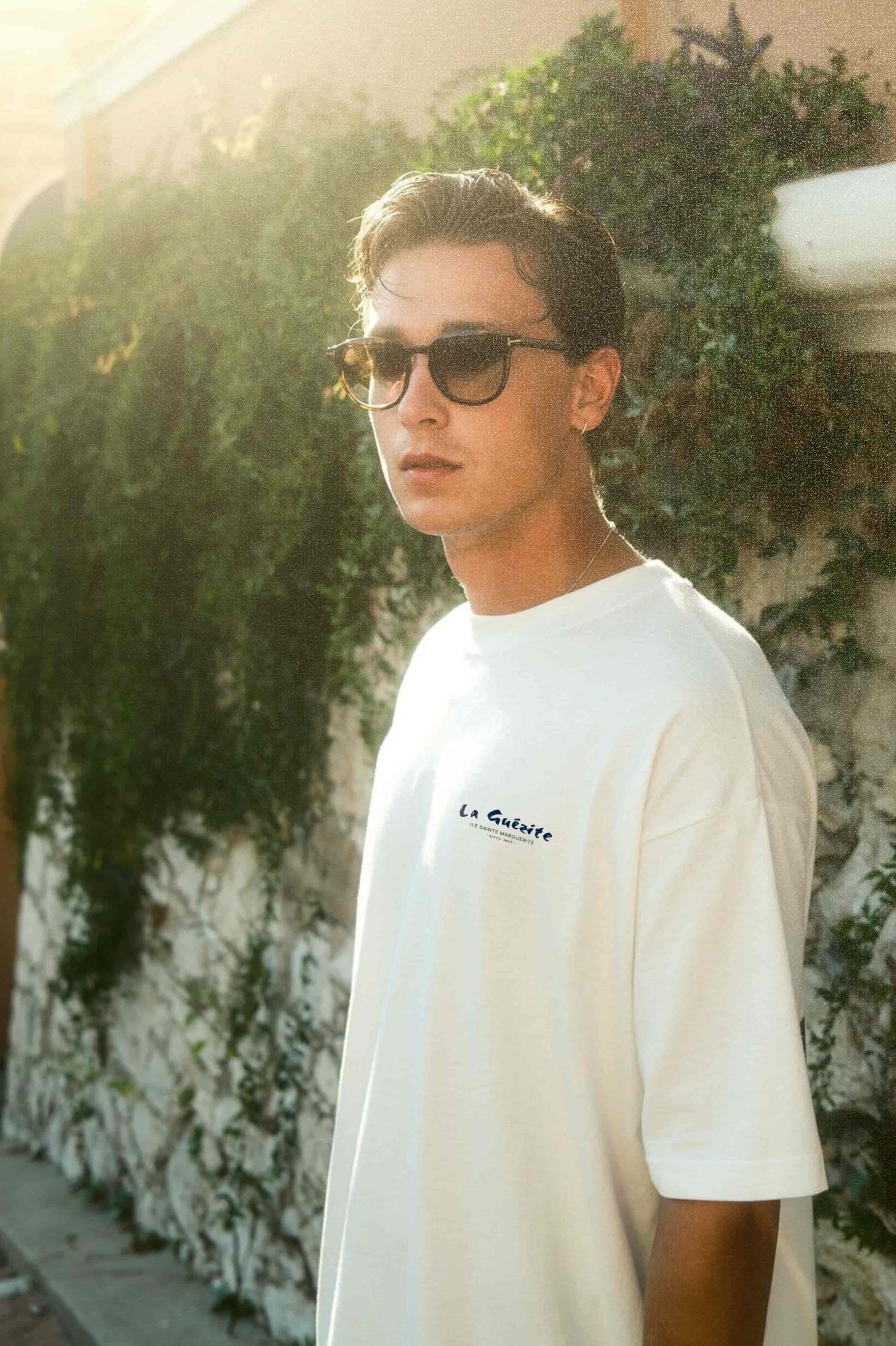 Man wearing sunglasses and a white t-shirt with a logo, standing in front of greenery.