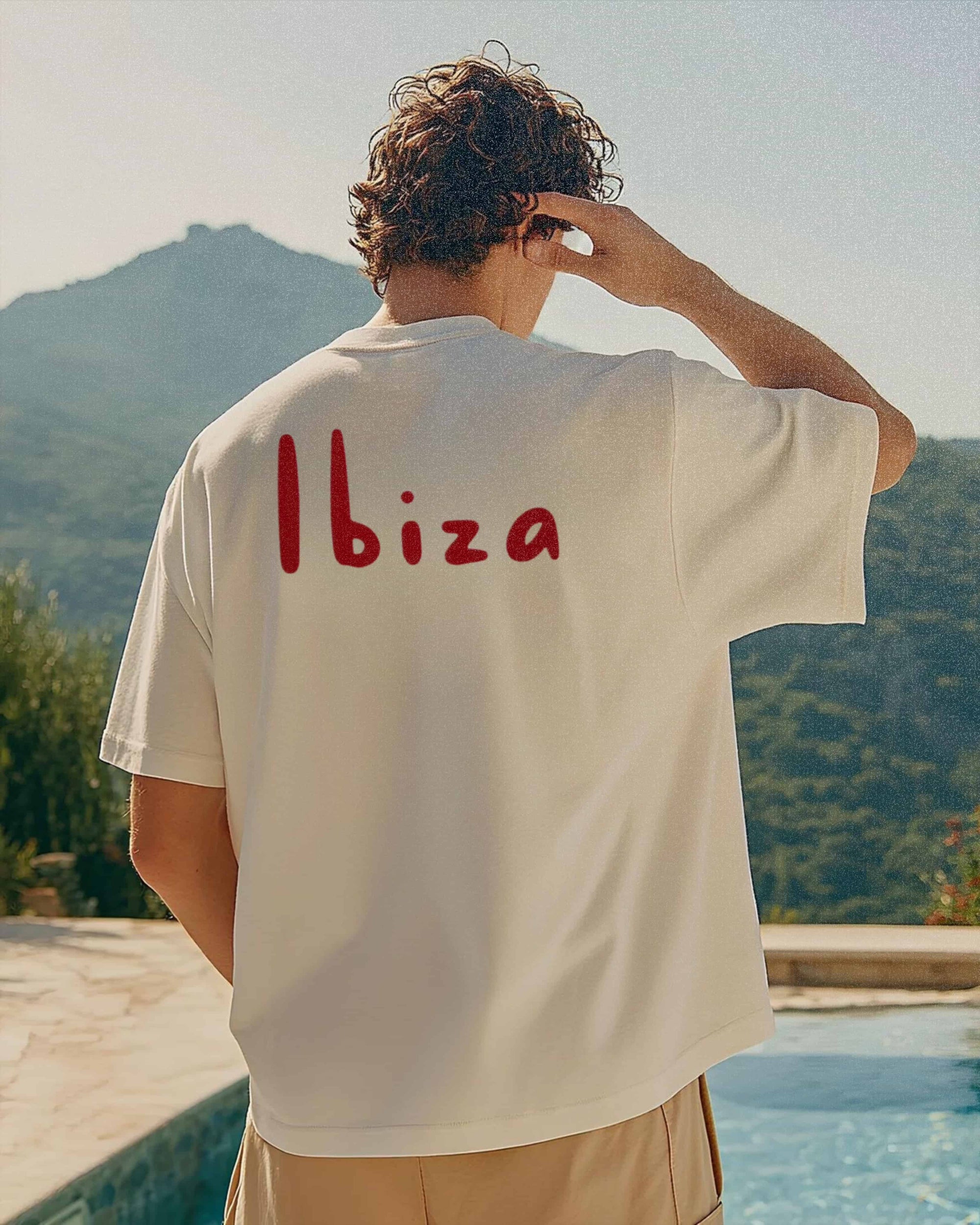 Person wearing a white t-shirt with 'Ibiza' on the back, standing by a pool with mountains in the background.