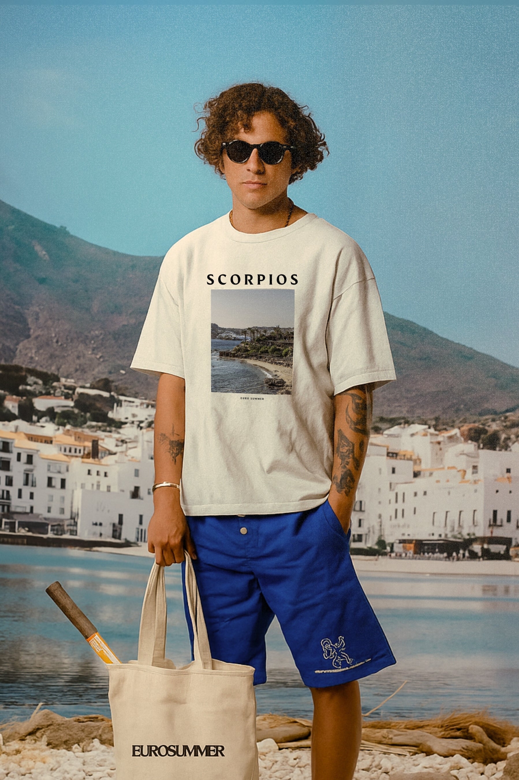 Person wearing a 'Scorpios' t-shirt with a scenic print, standing on a beach with mountains and buildings in the background.