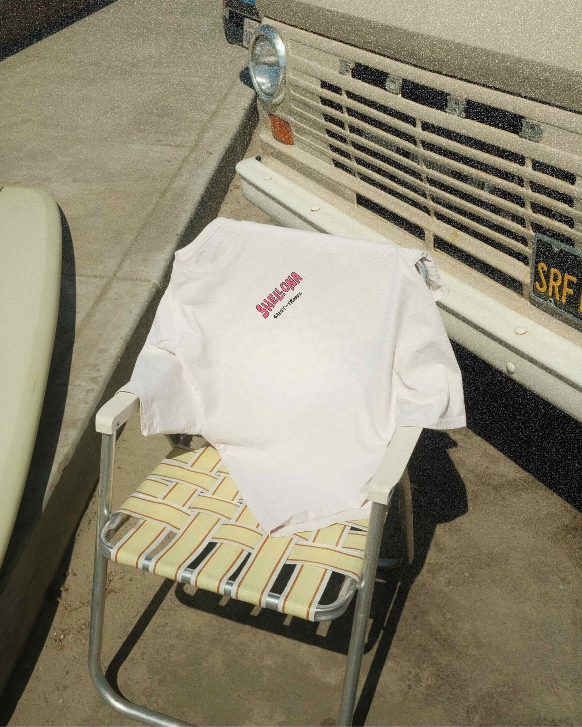 White t-shirt with red text on a yellow folding chair next to a vintage vehicle.